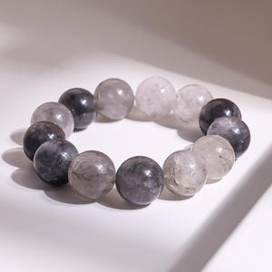 Könnte beinhalten: Ein Armband mit großen, runden Perlen in Grau-, Schwarz- und Klartönen. Die Perlen haben ein marmoriertes Aussehen und sind zu einer kreisförmigen Form aufgereiht. Das Armband wird auf einer weißen Oberfläche präsentiert.