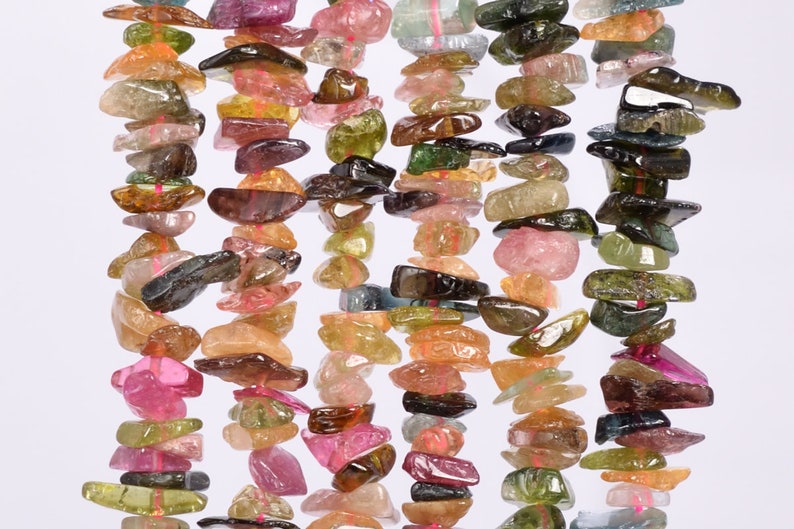 May include: Strands of colorful tourmaline gemstone chips in shades of pink, green, brown, and clear. The chips are irregularly shaped and strung together with pink thread, creating a textured and vibrant appearance. Ideal for jewelry making.