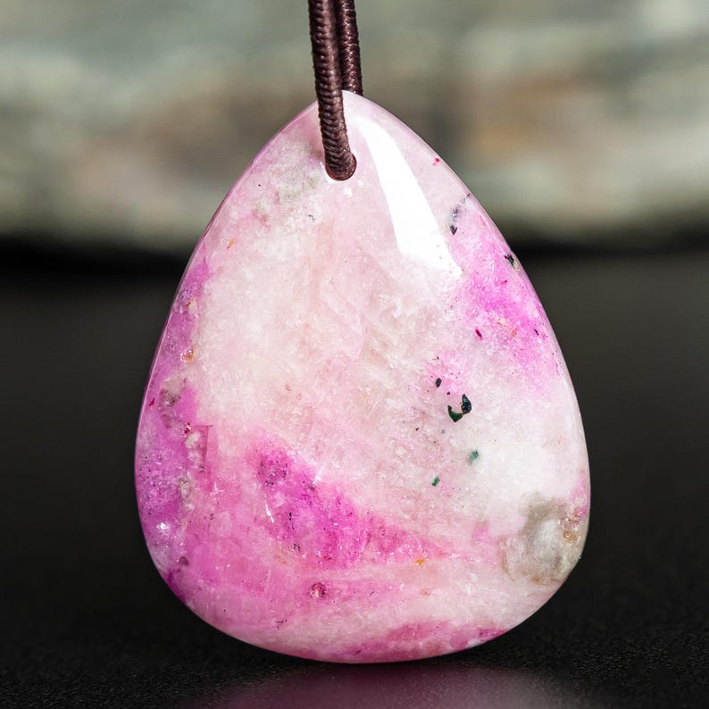 May include: A teardrop-shaped pendant made of polished stone with a mottled appearance, featuring vibrant shades of magenta and pink mixed with white and translucent crystalline areas, suspended from a dark brown cord through a drilled hole.