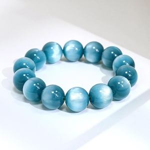May include: A stretch bracelet composed of smooth, round, light blue beads with a chatoyant, cat's eye effect. The beads are uniform in size and arranged in a circular loop, displaying a reflective, pearlescent sheen across their polished surfaces.