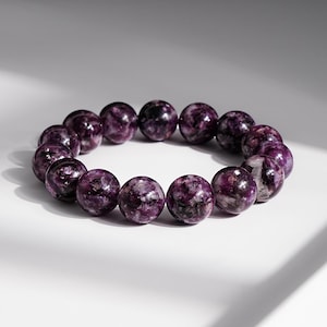 May include: A beaded bracelet featuring round, polished beads in shades of deep purple with white and black inclusions. The bracelet is displayed on a white surface, showcasing the natural variations in the gemstone.