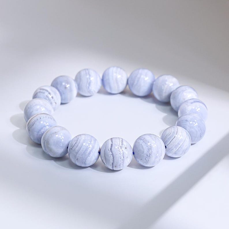May include: A light blue beaded bracelet. The bracelet features large, round beads with a marbled appearance. The beads are strung together to form a circular shape. The bracelet is set against a white background.