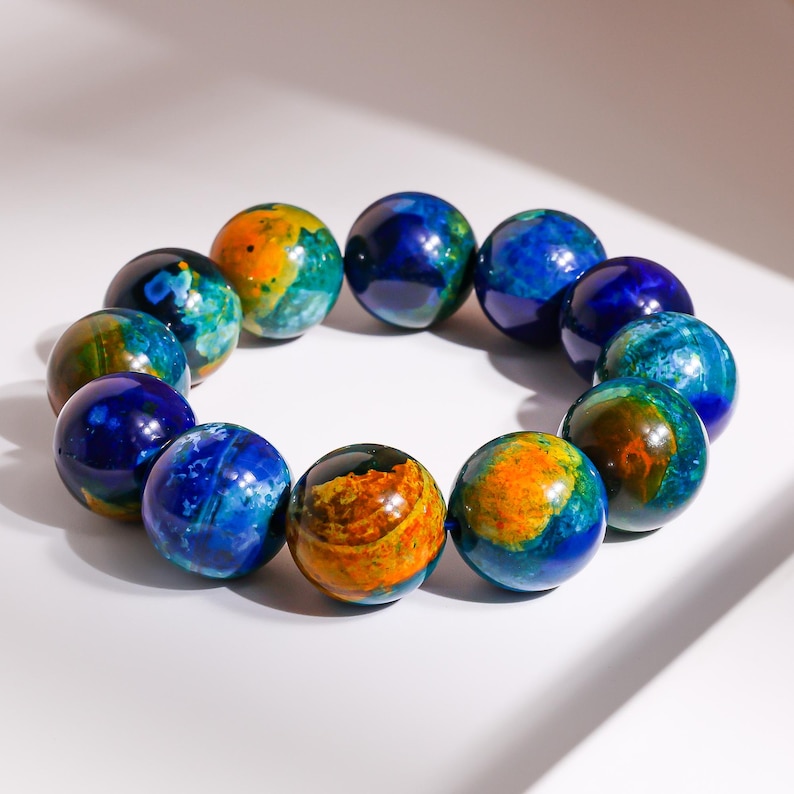 Pu&ograve; includere: Un braccialetto composto da dodici perline sferiche. Le perline sono principalmente blu, con vortici di arancione, giallo e verde. Le perline sono disposte in un motivo circolare, suggerendo un accessorio da polso.