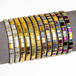 Könnte beinhalten: Eine Sammlung goldfarbener und farbenfroher Perlenarmbänder. Jedes Armband hat ein rechteckiges Kachelmuster mit verschiedenen Farben, darunter Gold, Weiß, Irisierend, Blau und Lila. Die Armbänder werden auf einer grauen Oberfläche präsentiert.