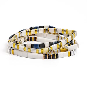 May include: A stack of four beaded bracelets. The bracelets feature rectangular beads in white, gold, silver, black, and navy blue. The bracelets are arranged in a circular pattern against a white background.