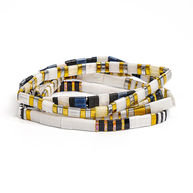 May include: A stack of four beaded bracelets. The bracelets feature rectangular beads in white, gold, silver, black, and navy blue. The bracelets are arranged in a circular pattern against a white background.