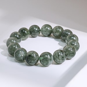 May include: A bracelet composed of round, translucent green beads with dark inclusions. The beads are strung together, forming a circular shape. The bracelet is displayed on a white surface.