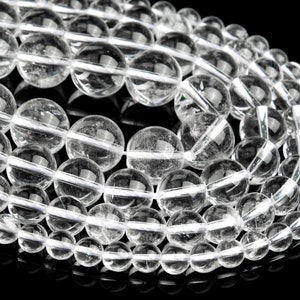 Puede incluir: Una hebra de cuentas de cuarzo transparente, cada cuenta es redonda y lisa. Las cuentas están dispuestas en un patrón circular, con los extremos de la hebra superpuestos.