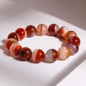 May include: A bracelet made of polished, round gemstone beads. The beads are a mix of red, brown, and cream colors, with variations in patterns and translucency. The bracelet is displayed on a white surface.