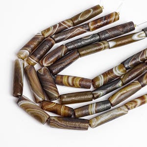 May include: A collection of cylindrical beads featuring natural banded patterns in shades of brown, tan, and cream. Each bead displays unique swirling layers and organic textures, strung together in a series to form a long, continuous strand.