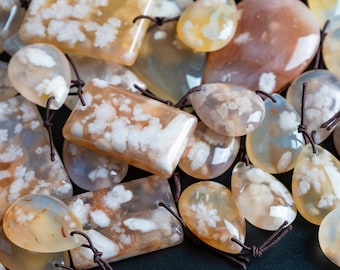 May include: A collection of polished flower agate pendants in various shapes, including rectangular and teardrop forms. Each translucent stone features creamy white, flower-like inclusions set against a warm, honey-colored base with dark brown cords.