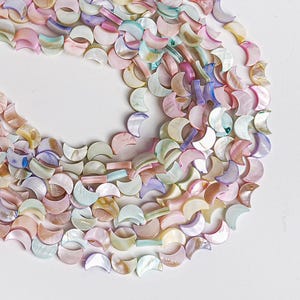 May include: A collection of iridescent, crescent-shaped beads in pastel colors including pink, blue, and yellow. The beads are scattered on a white surface, creating a visually appealing pattern. These beads are suitable for jewelry making.
