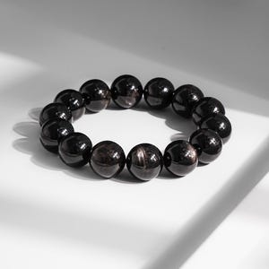 May include: A black beaded bracelet with a polished finish. The bracelet is composed of spherical beads, each displaying a subtle, reflective sheen. The beads are strung together, forming a circular shape. The bracelet is set against a white background.