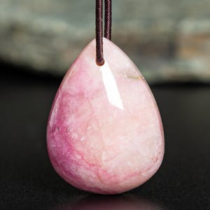 May include: A teardrop-shaped pendant made of polished pink stone with white crystalline inclusions. The pendant features a smooth, glossy surface and is suspended from a thin, dark brown cord that passes through a hole at the top of the stone.