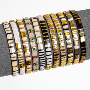 May include: A selection of ten beaded bracelets in diverse colours and designs. The bracelets feature gold, black, white, and other colourful beads. Some bracelets have stripes, while others have solid colours. The bracelets are displayed on a grey surface.