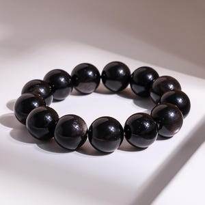 May include: A black beaded bracelet. The bracelet features large, round, polished black beads. The beads have a reflective, glossy surface. The bracelet is presented on a white surface with soft lighting.