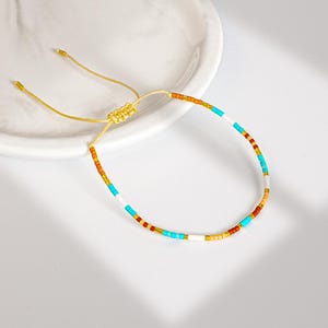 May include: A colorful beaded bracelet with a gold-colored adjustable cord. The bracelet features small, round beads in shades of orange, turquoise, white, and gold. The bracelet is displayed on a white surface.