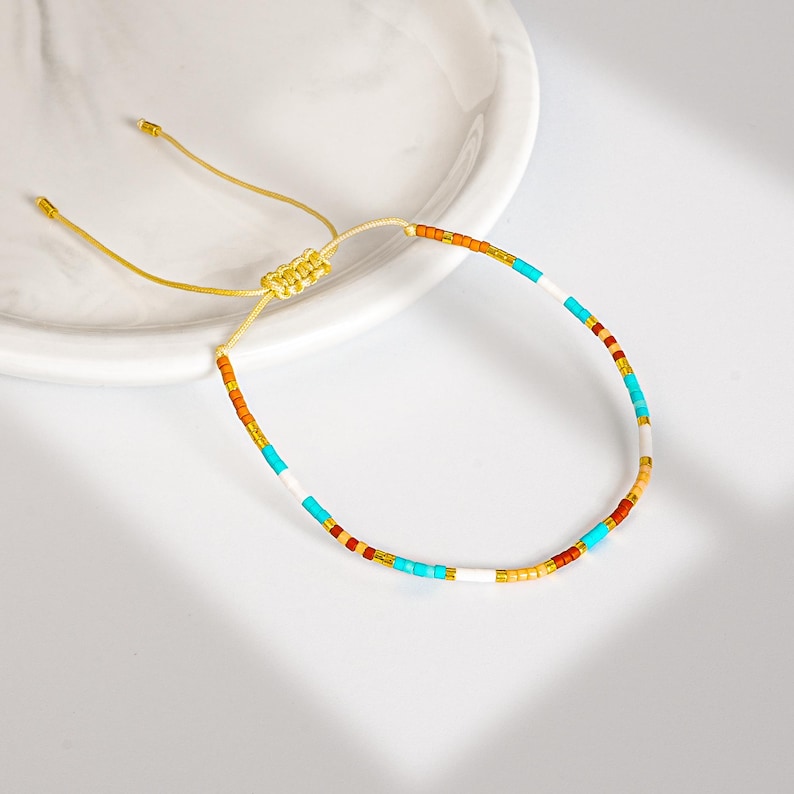 May include: A colorful beaded bracelet with a gold-colored adjustable cord. The bracelet features small, round beads in shades of orange, turquoise, white, and gold. The bracelet is displayed on a white surface.