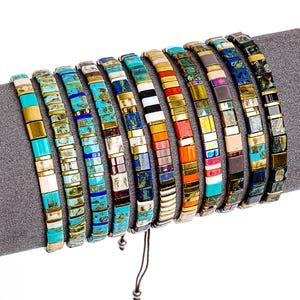 Może przedstawiać: Kolekcja kolorowych bransoletek z koralików. Każda bransoletka ma prostokątne koraliki w różnych kolorach, w tym turkusowym, złotym, czarnym, białym i pomarańczowym. Bransoletki są prezentowane na szarej powierzchni.