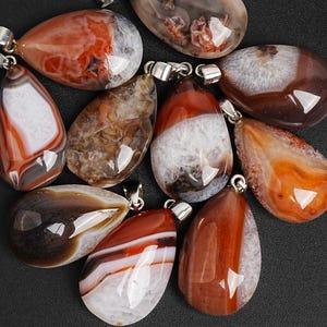 May include: A collection of polished agate pendants in various shapes and colors, including red, brown, white, and orange. Each pendant has a silver-colored bail for attaching to a chain or cord. The pendants are displayed against a dark background.