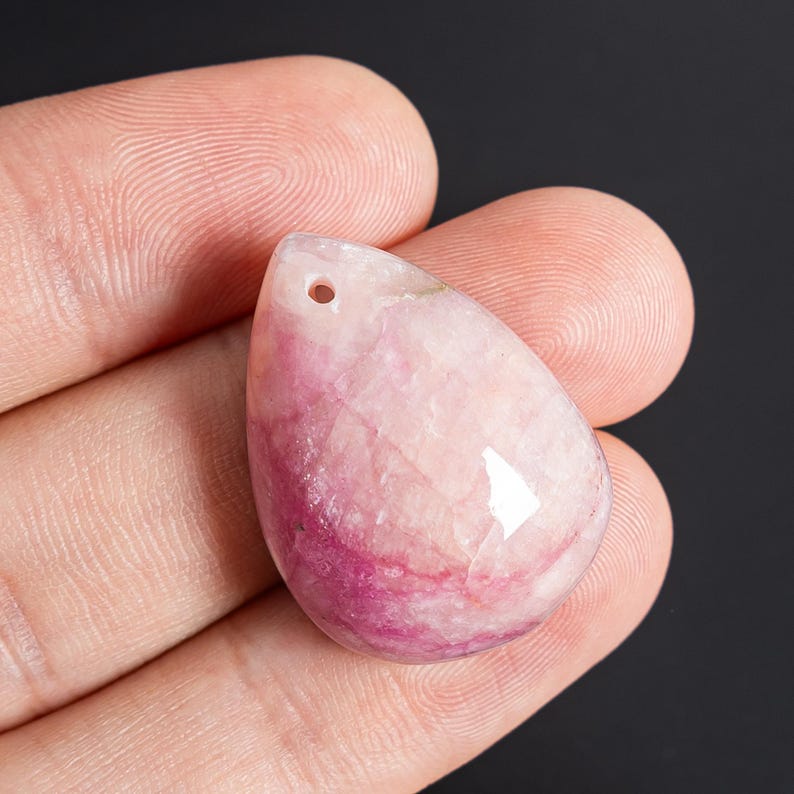 May include: A teardrop-shaped pendant made of polished stone with a smooth, glossy surface. The stone features a gradient of colors, transitioning from a deep, vibrant magenta at the base to a pale, translucent pink and white at the top.
