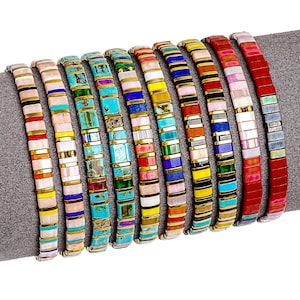 May include: A collection of colorful beaded bracelets. Each bracelet features a unique pattern of rectangular beads in various colors, including turquoise, pink, yellow, and red, separated by gold-colored metal accents. The bracelets are displayed on a gray surface.
