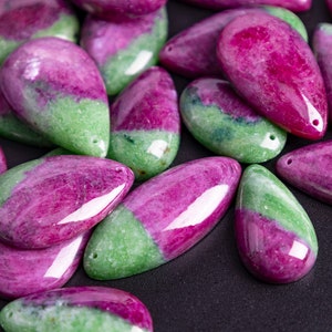 May include: A selection of polished gemstone pendants. Each pendant is a teardrop shape, with a vibrant mix of colours. The stones are a combination of deep magenta and light green, with a hole drilled at the top for stringing.