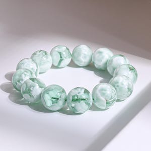 Könnte beinhalten: Ein Armband mit großen, runden Perlen in Hellgrün und Weiß. Die Perlen haben ein marmoriertes Aussehen mit transluzenter Qualität. Das Armband ist aufgefädelt und bildet eine kreisförmige Form. Die Perlen haben einen Durchmesser von etwa 2,5 cm.