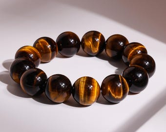 Puede incluir: Un brazalete hecho de cuentas de piedra preciosa ojo de tigre pulidas. Las cuentas redondas presentan una mezcla de colores marrón y amarillo dorado. El brazalete se muestra sobre una superficie blanca.