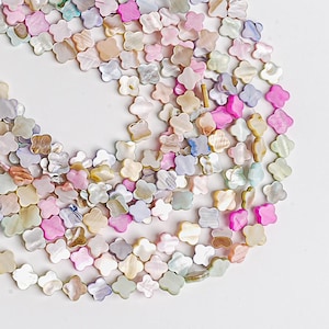 May include: A collection of iridescent, four-petal flower-shaped beads in various pastel colors, including pink, white, beige, and green. The beads are scattered on a white surface, creating a visually appealing pattern. These beads are ideal for jewelry making.