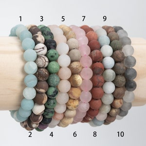 May include: Ten bracelets made of different types of stones, including green, brown, white, pink, and gray. The bracelets are arranged in a row on a wooden stand, with numbers 1 through 10 labeling each bracelet.