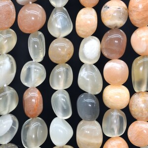 May include: A strand of smooth, oval-shaped sunstone beads in shades of peach, white, and light orange. The beads have a translucent quality and show a shimmery effect.