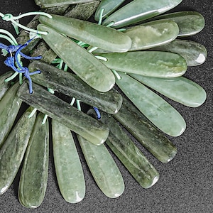 May include: A collection of teardrop-shaped jade pendants in varying shades of green. Each pendant is strung with green and blue cord, suggesting they are intended for jewelry making or crafting. The polished stones have natural veining.