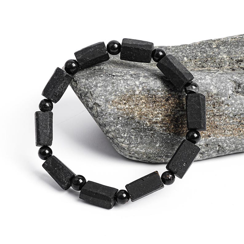 May include: A black beaded bracelet featuring rectangular and round beads. The bracelet is made of dark, possibly natural, stones and is displayed on a gray rock against a white background. The bracelet is a piece of jewelry.