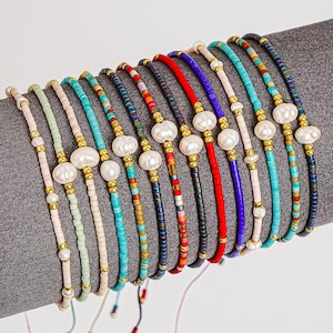 May include: A collection of beaded bracelets in various colors, including turquoise, red, and navy. Each bracelet features a central pearl accent and gold-colored beads. The bracelets are displayed on a gray surface.