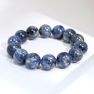 May include: A stretch bracelet composed of round, polished kyanite beads. The beads feature a deep blue color with natural variations, including streaks of lighter blue, grey, and dark inclusions, creating a marbled appearance throughout the piece.