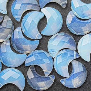 Pode incluir: Uma coleção de cabochões de vidro em forma de lua crescente azul iridescente. Os cabochões têm um design facetado e geométrico.