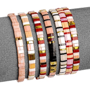 May include: A collection of seven beaded bracelets in various colors, including pink, white, red, gold, and black. The bracelets are made of small, flat beads and are displayed on a gray surface. Each bracelet has a different color scheme.