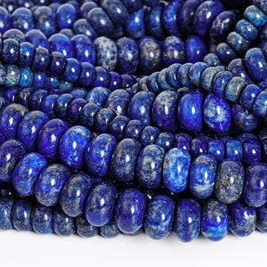 May include: Close-up of strands of polished, oval-shaped lapis lazuli beads. The beads are a deep, rich blue color with flecks of gold and white, creating a textured appearance. The beads are strung together, suggesting they are for jewelry making.