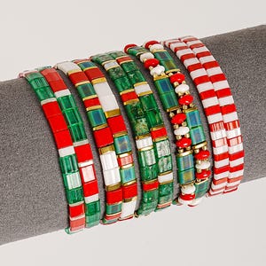 May include: A collection of beaded bracelets in festive colors. The bracelets feature red, green, white, and gold beads, arranged in various patterns, including stripes and blocks. The bracelets are displayed on a gray surface.
