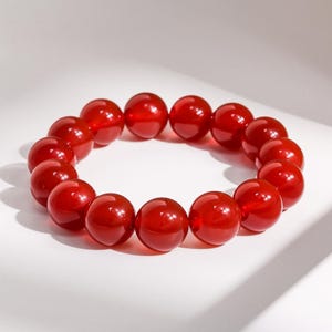 May include: A bracelet made of polished, round, red beads. The beads are strung together to form a circular shape. The beads have a glossy finish and reflect light, creating a vibrant appearance. The bracelet is displayed on a white surface.
