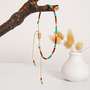 May include: A colorful beaded necklace with a sliding knot closure. The necklace features turquoise, red, orange, yellow, black, and gold beads. The necklace is displayed on a small, brown branch against a white background.