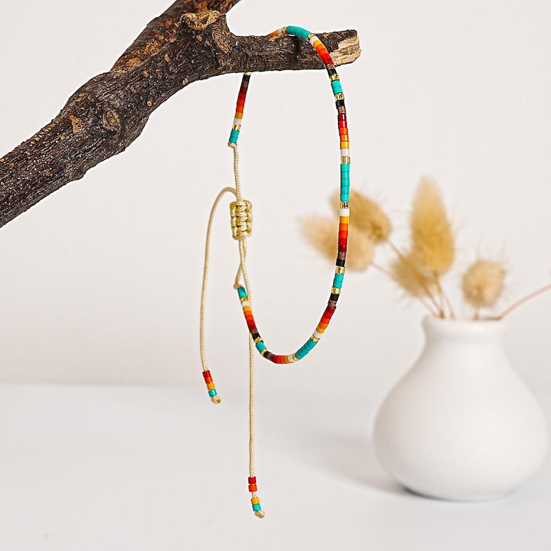 May include: A colorful beaded necklace with a sliding knot closure. The necklace features turquoise, red, orange, yellow, black, and gold beads. The necklace is displayed on a small, brown branch against a white background.