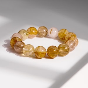 May include: A beaded bracelet featuring a series of translucent, round beads in shades of gold, amber, and pale pink. The beads are strung together, forming a circular shape. The bracelet is displayed on a white surface.
