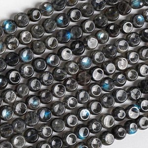 May include: A collection of round, dark gray labradorite beads arranged in rows. Each bead features a smooth, polished surface with natural inclusions and iridescent blue flashes that catch the light, creating a reflective and textured appearance.