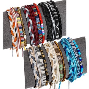 Puede incluir: Una colección de pulseras tejidas y con cuentas en varios colores, incluyendo naranja, blanco, rojo, negro y azul. Algunas pulseras presentan diseños trenzados, mientras que otras tienen cuentas coloridas. Las pulseras se exhiben sobre una superficie gris.
