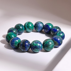Peut inclure: Un bracelet composé de perles sphériques polies. Les perles présentent un mélange de bleu profond et de vert, avec un aspect marbré. Le bracelet est présenté sur une surface blanche, avec des ombres douces.