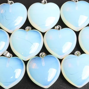 May include: A collection of heart-shaped pendants with a pearlescent, opalescent appearance. Each pendant has a silver-colored bail for attaching to a chain. The hearts have a soft, light blue hue with a subtle, swirling internal pattern.