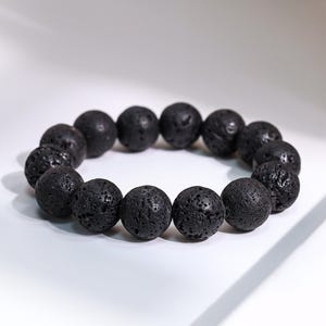 Peut inclure: Un bracelet de perles en pierre de lave noire. Le bracelet présente un design circulaire de perles sphériques poreuses. Les perles sont d'une couleur charbon foncé et ont une surface texturée. Le bracelet est présenté sur une surface blanche.