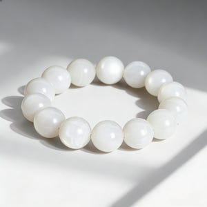 May include: A white beaded bracelet with large, round beads. The bracelet is made of smooth, polished beads, and is displayed on a white surface. The beads have a slight sheen, reflecting the light.